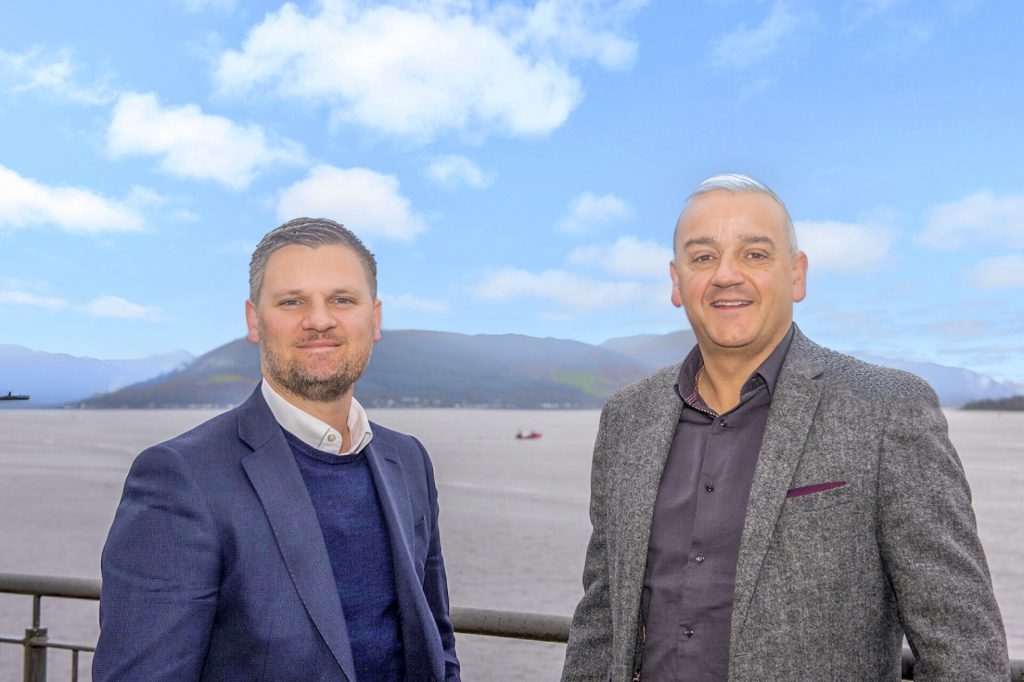 picture of two men in jackets standing in front of water and hills